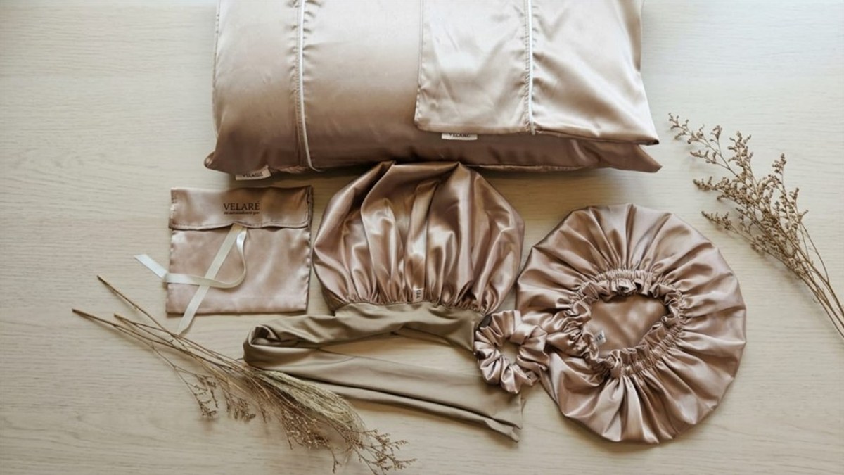 Champagne satin VELARÉ pillowcase, bonnets, scrunchie, and pouch arranged on wood